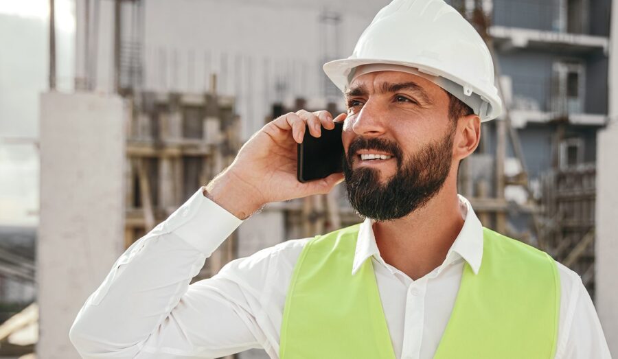 Busy bearded male architect in hardhat and waistcoat on phone at construction site getting advice on construction business growth training