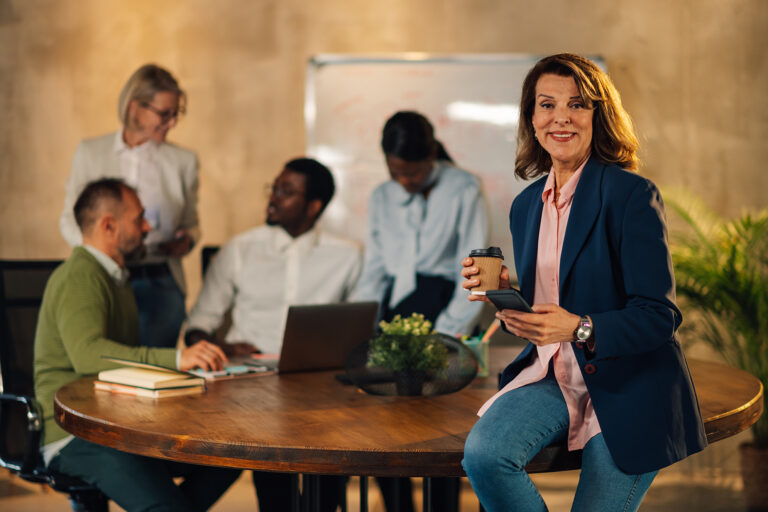 Smiling woman holding a coffee cup and phone with a collaborative team meeting happening behind her, representing positive workplace wellbeing.